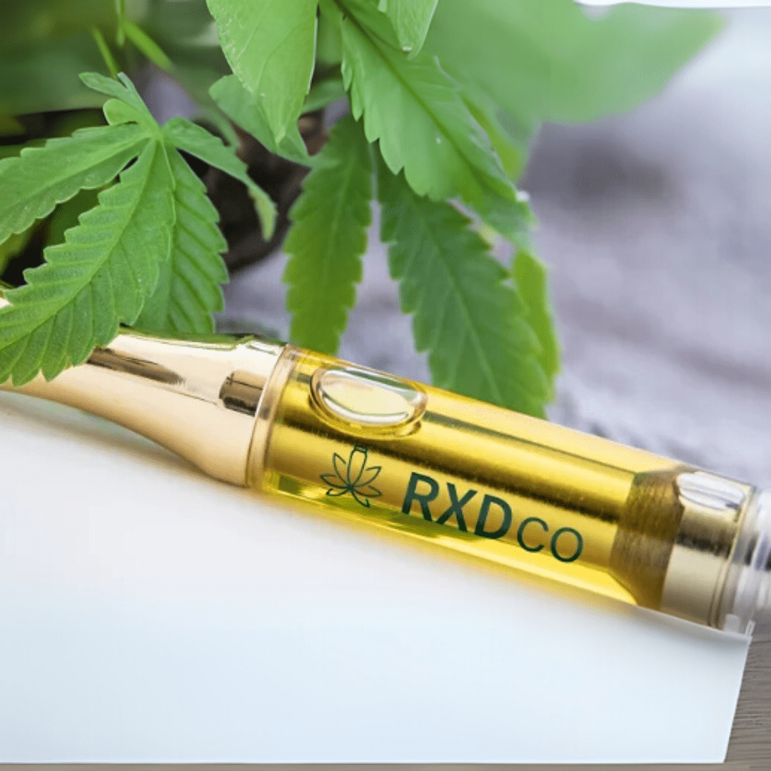 Gold ceramic vape cartridge filled with cannabis oil, branded with RXDco logo, placed in front of cannabis leaves.